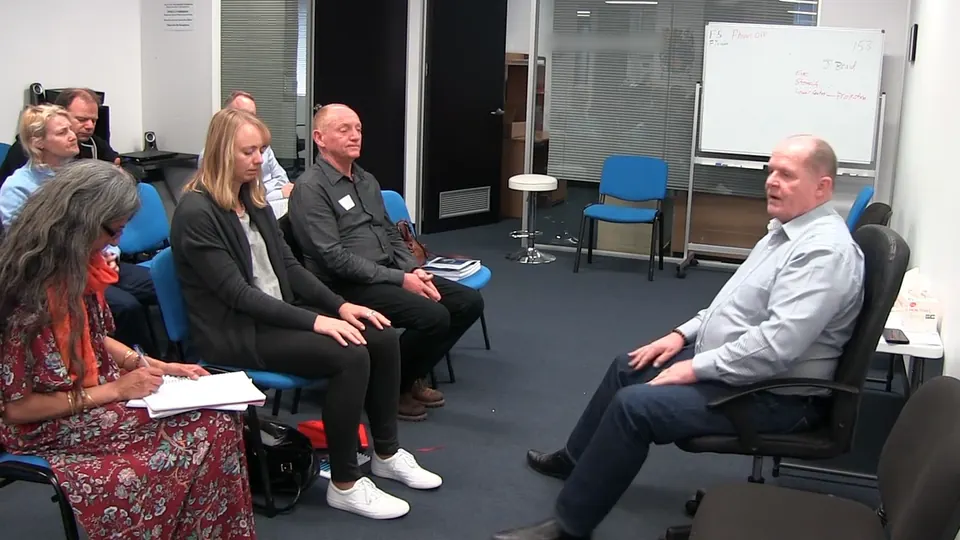 Rick Collingwood seated facing a row of students, one writing in a notebook, with a whiteboard behind them