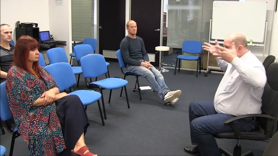 Rick Collingwood seated with both hands raised, explaining to two students in a room of blue chairs