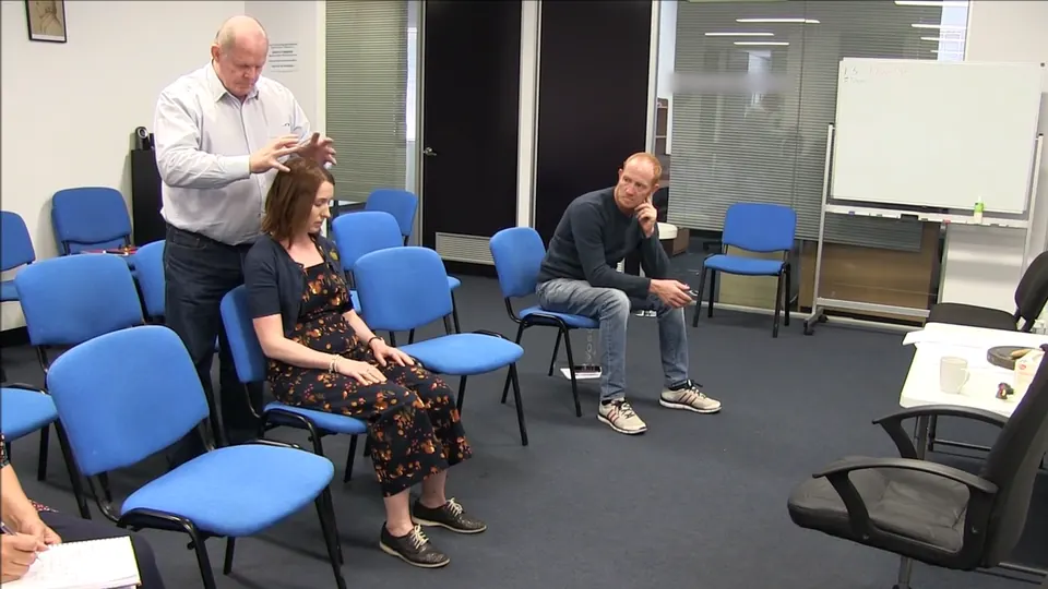Rick Collingwood standing behind a seated woman with his hands above her head, while a student watches