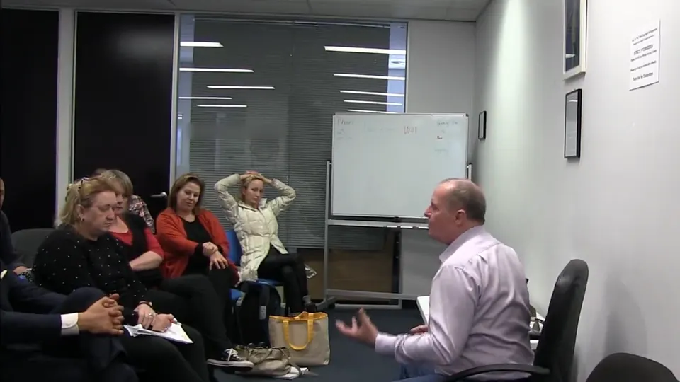 Rick Collingwood seated at the front of a training room, gesturing to a group of students with a whiteboard behind