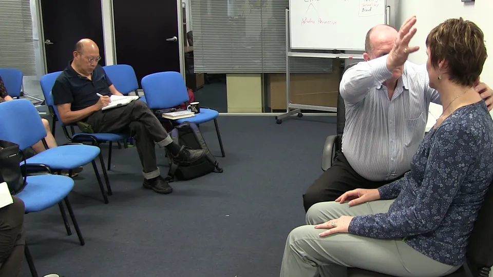 Rick Collingwood seated beside a woman with one arm raised, while a student writes in a notebook