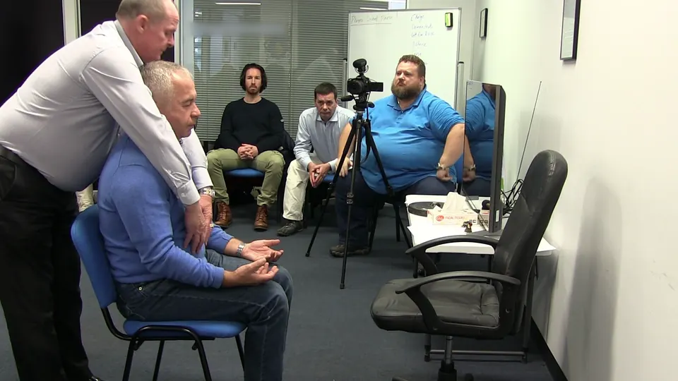 Rick Collingwood leaning over a seated man during a demonstration, watched by three students and a camera on a tripod