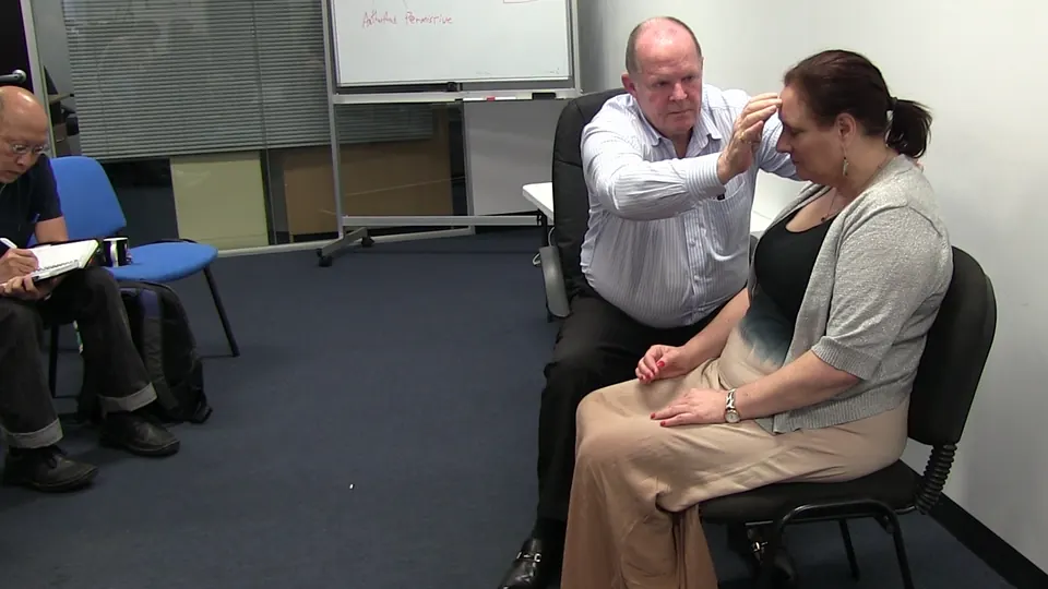 Rick Collingwood seated beside a woman with his fingers on her forehead, while a student takes notes