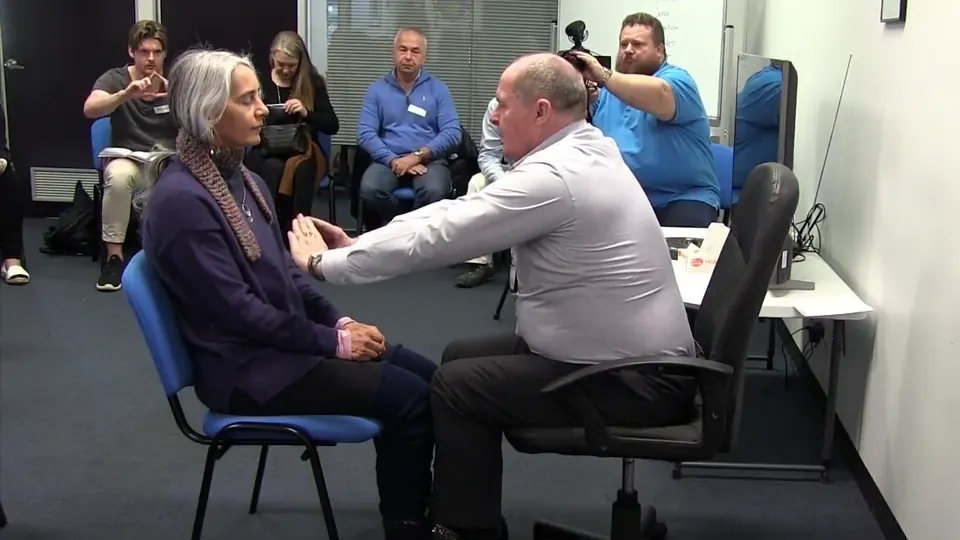 Rick Collingwood seated facing a student with his hands raised in a mesmeric induction, as the class watches
