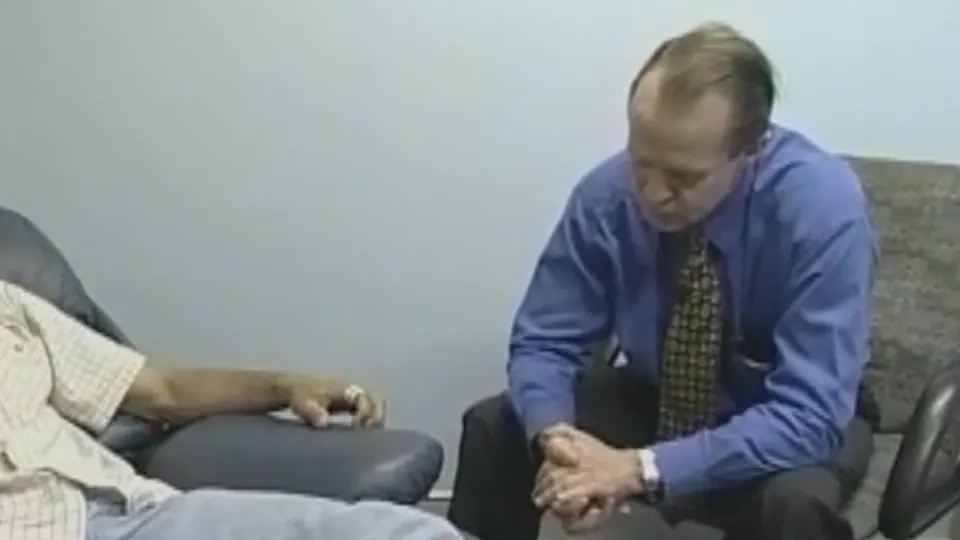 The hypnotherapist seated with hands clasped beside a recliner during a recorded clinical hypnotherapy session